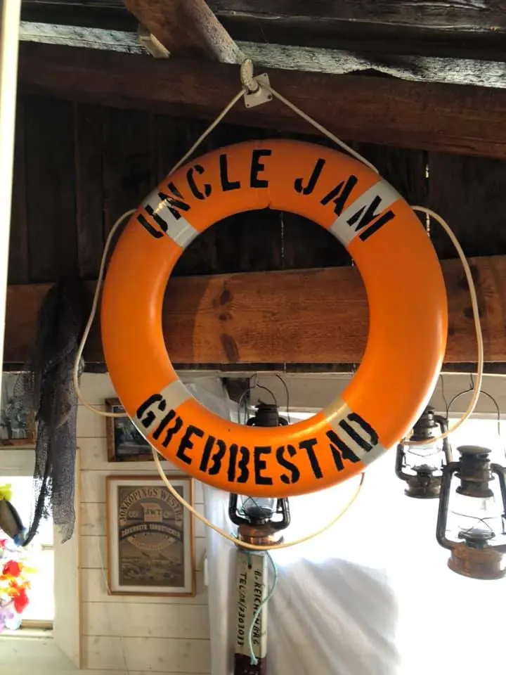 Orange livboj med svart text "Uncle Jam Grebbestad" hängande i en rustik träbyggnad.